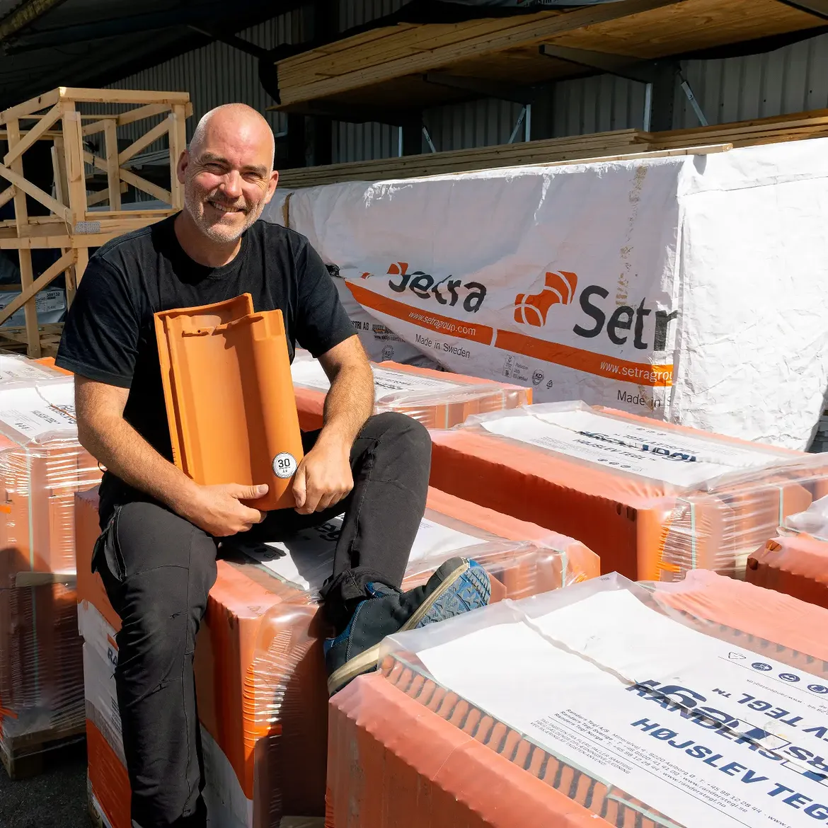 Leende medarbetare i lagret som håller upp en orange tegelpanna vid pallar med taktegel